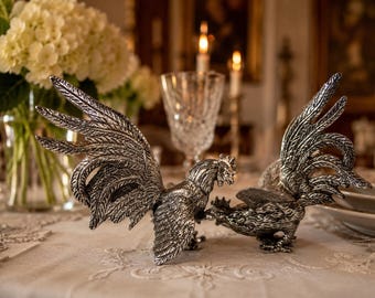 May include: Two silver rooster figurines with detailed feather designs are displayed on a table. The roosters are positioned facing each other, showcasing their intricate craftsmanship. The background includes a vase of white flowers and lit candles.