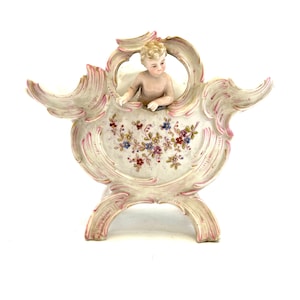 May include: A vintage porcelain figurine featuring a cherub figure within a decorative, ornate frame. The frame is primarily off-white with pink and gold accents, and floral detailing. The cherub is nude with blonde hair. The figurine has a unique, antique aesthetic.