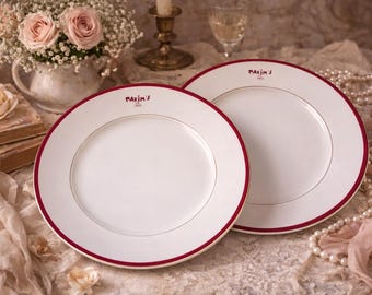 May include: Two white ceramic plates with a red border and the text "Maxim's Paris" in red. The plates are on a lace tablecloth, with flowers and pearls in the background. The plates are for serving food.