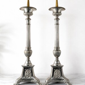 May include: A pair of antique silver-toned candlesticks. Each candlestick features a fluted column, ornate base with a decorative emblem, and a gold-colored candle holder. The candlesticks are displayed on a marble surface.