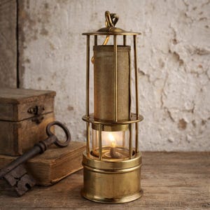 Op de afbeelding: Een messing mijnwerkerslamp met een brandende kaars. De lamp heeft een gaasscherm en een glazen gedeelte. Hij staat op een houten oppervlak, met een sleutel en een kleine houten kist op de achtergrond. Vintage design.