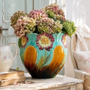 Op de afbeelding: Een turquoise keramische vaas met een bloemenontwerp, gevuld met roze en groene hortensia's. De vaas heeft grote, gestileerde bloemen in tinten geel, bordeauxrood en bruin. De vaas staat op een witte houten tafel.