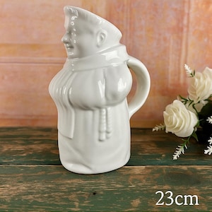 May include: White antique monk pitcher, French Barbotine, with a handle and a detailed face. The pitcher is 9 inches tall and sits on a wooden surface. The text "Antique Monk Pitcher French Barbotine" is displayed.