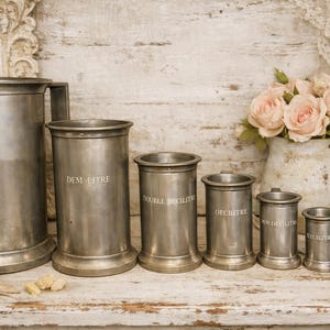 Op de afbeelding: Een verzameling antieke metalen maatbekers en een kan met roze rozen. De maatbekers zijn gemarkeerd met Franse volume-eenheden, waaronder "UN LITRE" en "DEM LITRE". De items zijn gerangschikt op een houten oppervlak.