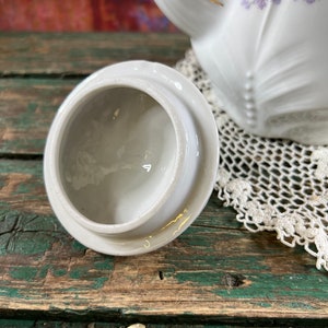 May include: White porcelain teapot lid with a floral design. The lid is round and has a small knob on top. The lid is sitting on a wooden surface.