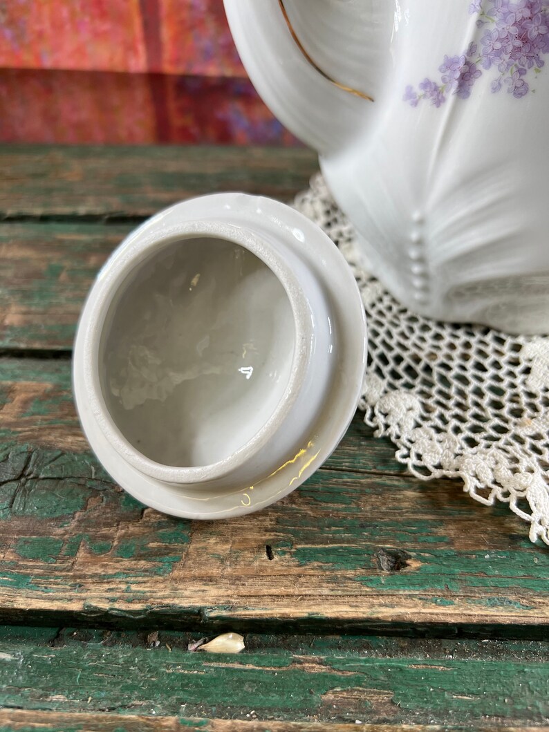 May include: White porcelain teapot lid with a floral design. The lid is round and has a small knob on top. The lid is sitting on a wooden surface.