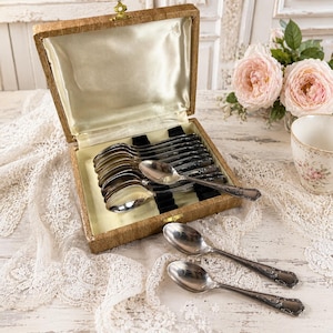 May include: A set of silver-toned metal spoons with ornate embossed handles, arranged inside a rectangular box with a textured tan exterior and a cream-coloured satin lining. Two additional spoons rest outside the box on a white lace fabric.