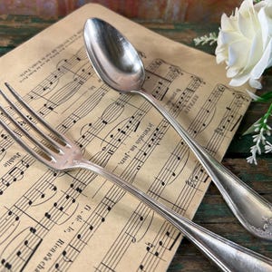 May include: A silver fork and spoon set on a sheet of music. The fork has four tines and the spoon has a rounded bowl. Both pieces are polished and shiny.