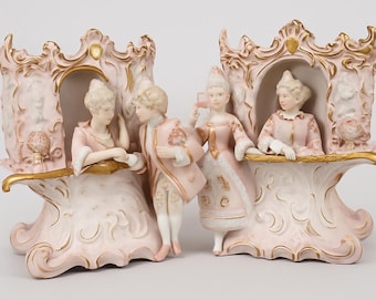 May include: Two porcelain figurines depicting a scene with figures in ornate carriages. The figurines are primarily pink and white with gold accents. The figures are dressed in period clothing, suggesting a historical or romantic theme.