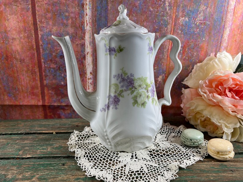 May include: A white porcelain teapot with a floral design featuring purple and green flowers. The teapot has a delicate, ornate handle and a lid with a finial. The teapot is sitting on a white crocheted doily.