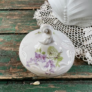 May include: White porcelain teapot lid with a floral design featuring purple and green flowers. The lid has a knob on top and a gold trim around the edge.