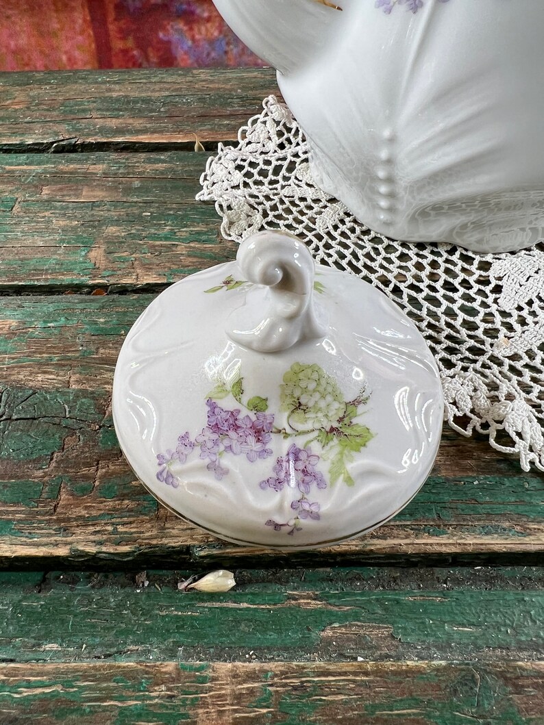 May include: White porcelain teapot lid with a floral design featuring purple and green flowers. The lid has a knob on top and a gold trim around the edge.