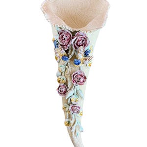 May include: Cream-colored ceramic wall pocket vase shaped like a cornucopia, adorned with pink roses, blue and yellow birds, and green leaves. The top has a decorative pattern and a small hole for hanging. A decorative floral accent.