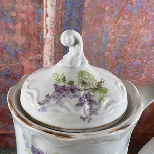 May include: White porcelain teapot with a floral design. The teapot has a lid with a floral design and a gold trim. The teapot is decorated with purple and green flowers.