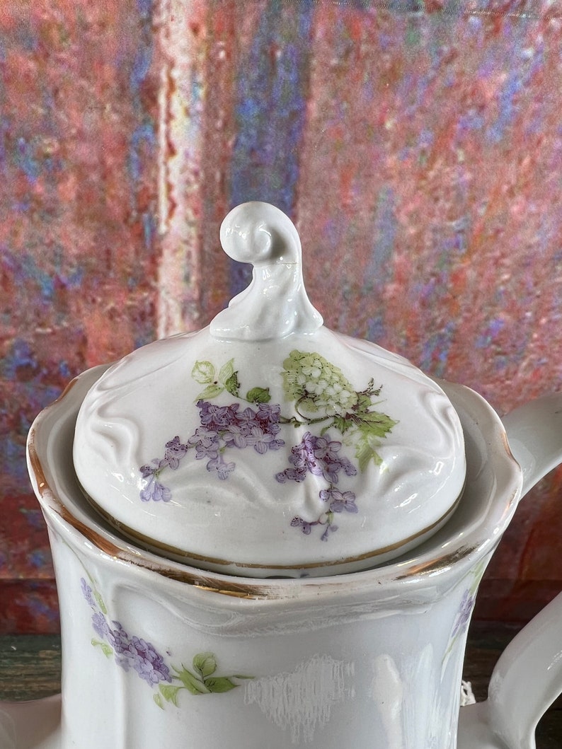 May include: White porcelain teapot with a floral design. The teapot has a lid with a floral design and a gold trim. The teapot is decorated with purple and green flowers.
