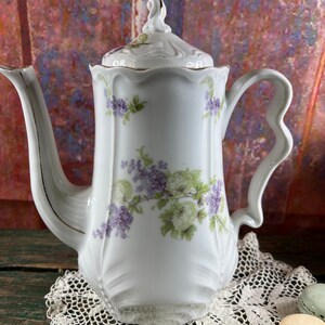 May include: White porcelain teapot with a floral design featuring purple and green flowers. The teapot has a gold trim around the lid and spout.