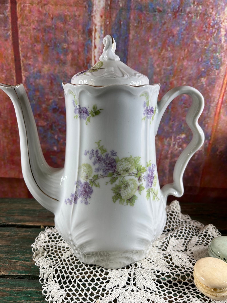 May include: White porcelain teapot with a floral design featuring purple and green flowers. The teapot has a gold trim around the lid and spout.