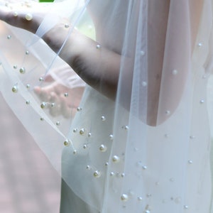 May include: A white tulle veil with scattered pearl embellishments. The veil is sheer and delicate, with a soft, flowing texture.