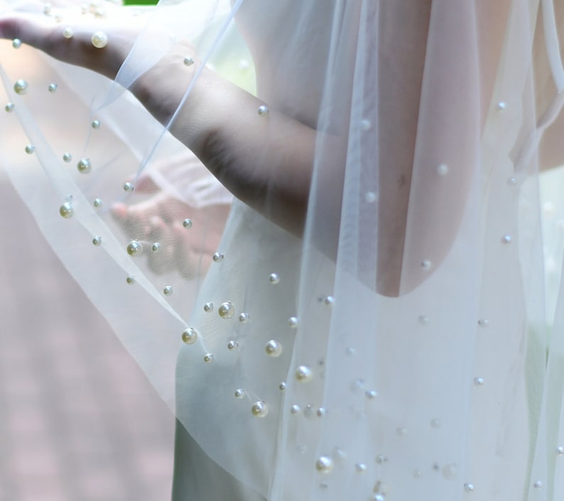May include: A white tulle veil with scattered pearl embellishments. The veil is sheer and delicate, with a soft, flowing texture.