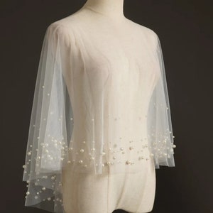 May include: A white tulle bridal cape with pearl embellishments. The cape has a round neckline and long sleeves. The pearls are scattered throughout the cape, creating a delicate and elegant look.