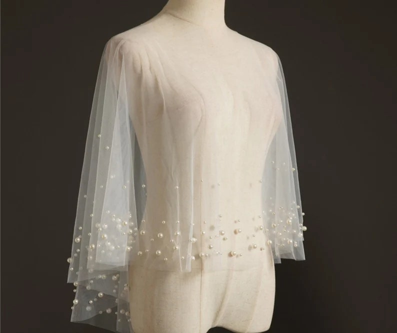 May include: A white tulle bridal cape with pearl embellishments. The cape has a round neckline and long sleeves. The pearls are scattered throughout the cape, creating a delicate and elegant look.