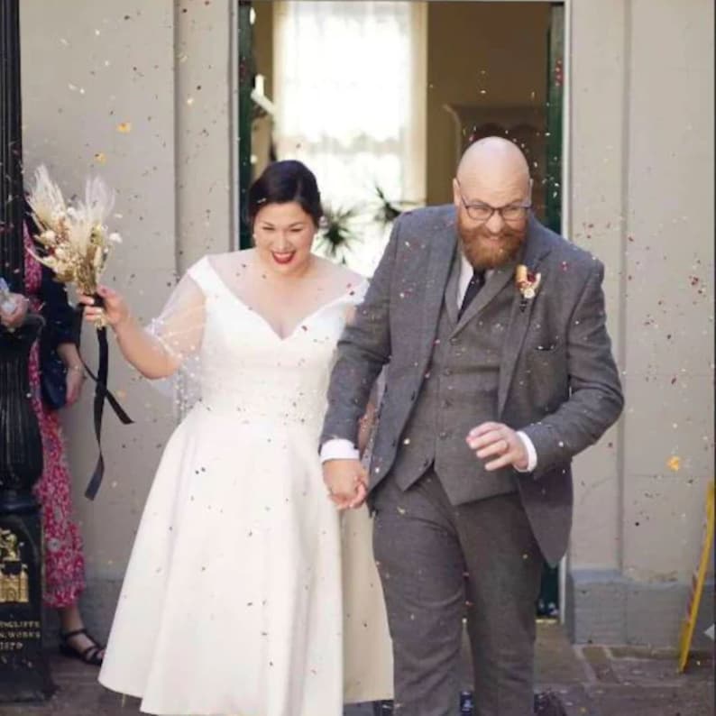 May include: A bride and groom walking out of a building, the bride is wearing a white dress and holding a bouquet of dried flowers, the groom is wearing a gray suit and a tie, confetti is falling around them.