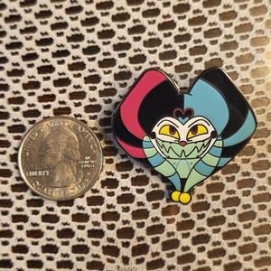 May include: A colourful enamel pin depicting a cartoon character with a heart-shaped head. The pin has a black background with pink and blue accents. The character has yellow eyes and a green body. A coin is next to the pin.