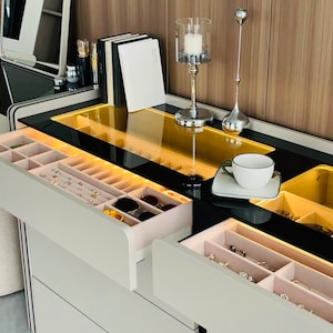 May include: A white vanity with a black glass top and drawers. The vanity has a built-in light and a glass top. The drawers are open and contain jewelry, sunglasses, and other accessories.