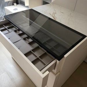 Könnte beinhalten: Eine weiße Schublade mit einer Glasplatte, die geöffnet ist und ein graues, mit Filz ausgekleidetes Fach mit mehreren Fächern zur Aufbewahrung von Schmuck zeigt.