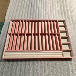 May include: A pink jewelry organizer with multiple compartments. The organizer has vertical slots, ring holders, and small square compartments. The interior is a light beige color, and the exterior is a soft pink.