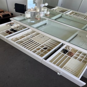 May include: A white jewelry organizer with multiple drawers, each sectioned to hold various jewelry pieces. The drawers are open, revealing necklaces, bracelets, rings, and other accessories. The organizer has a glass top and is set against a neutral background.