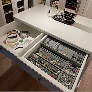 May include: White vanity table with two drawers, one open to reveal a variety of headbands and hair accessories, and the other open to reveal a variety of earrings, necklaces, and other jewelry. The table has a gray top and is made of wood.