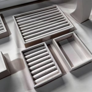 May include: A collection of white jewelry display trays. The trays are rectangular with a soft, gray interior designed to hold rings and other small jewelry items. The trays are arranged on a white surface.