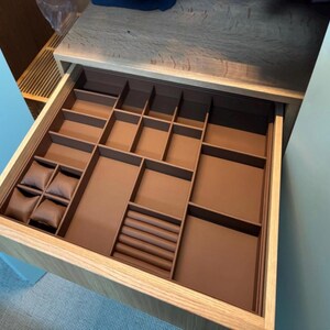 May include: Open wooden drawer with a brown interior organizer. The organizer has various compartments, including small square sections, rectangular spaces, and ring holders. Several small, brown, square pillows are visible in the left corner. The drawer is set in a light-colored wooden cabinet.
