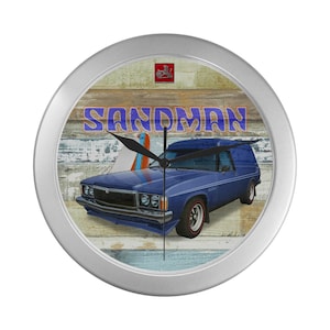 May include: A silver wall clock with a blue and white woodgrain background. The clock features a blue Holden panel van with surfboards and the text "SANDMAN".