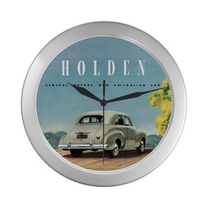 May include: A silver wall clock featuring a vintage advertisement for a Holden car. The ad shows a light grey Holden car with the text "Holden, General Motors New Australian Car".