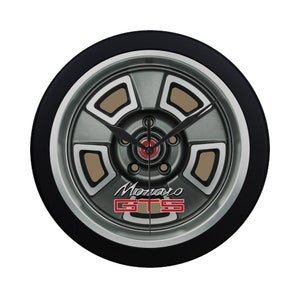 May include: A black and silver wall clock with a car wheel design. The clock face features a grey wheel with brown accents and the text "Monaro GTS" in red and silver lettering.