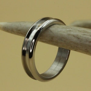 May include: A polished silver ring with a grooved design. The ring is displayed on a light-colored antler, set against a neutral background. The ring's reflective surface catches the light, highlighting its metallic sheen.