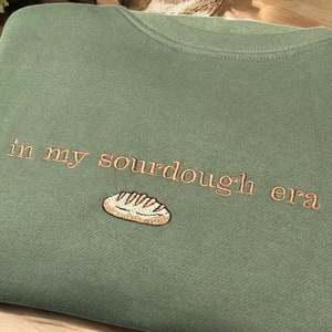 May include: Sage green sweatshirt with the embroidered phrase "in my sourdough era" and a bread loaf graphic. The text and graphic are in a light brown color. The sweatshirt is folded on a wooden surface.