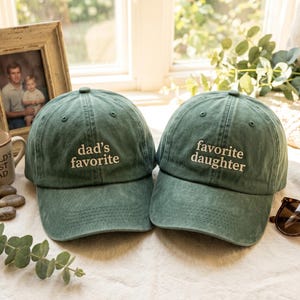 May include: Two green baseball caps with white embroidered text. One says "dad's favorite" and the other says "favorite daughter." The caps are on a white surface, with a framed photo and a mug in the background.