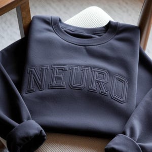 May include: A dark grey sweatshirt featuring the embroidered word "NEURO" across the chest. The sleeves are cuffed. The sweatshirt is folded on a chair. The fabric appears soft and comfortable.