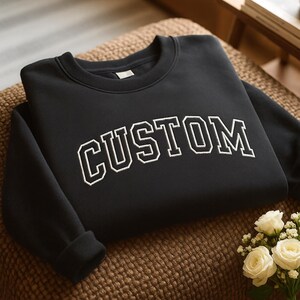 May include: Black sweatshirt with the word "CUSTOM" in white, block-style lettering. The sweatshirt is folded on a brown, textured surface. A small bouquet of white roses sits nearby.