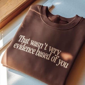 May include: A brown sweatshirt with the embroidered text "That wasn't very evidence based of you." The crew neck sweatshirt is made of a soft material and is folded on a white surface.