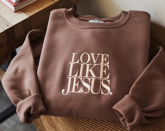 May include: A brown sweatshirt with the embroidered text "LOVE LIKE JESUS" in cream. The sweatshirt is folded on a brown corduroy surface. The sleeves are cuffed.
