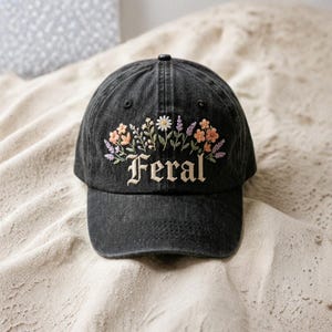 May include: Black denim baseball cap with embroidered floral design in shades of peach, lavender, and white. The word "Feral" is embroidered in a decorative font below the flowers. The cap has a curved brim and a button on top.