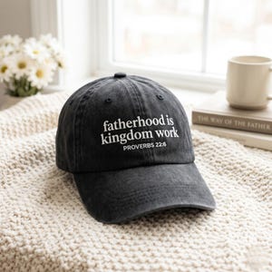 May include: A dark gray baseball cap with the text "fatherhood is kingdom work" and "PROVERBS 22:6" in white. The cap is made of a soft, worn material and has a curved brim. The hat is sitting on a cream-colored knitted blanket.