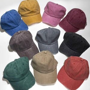 May include: A collection of ten baseball caps in various colors, including blue, yellow, pink, burgundy, black, and green. Each cap has a curved brim and a fabric-covered button on top. The caps are arranged on a white surface.
