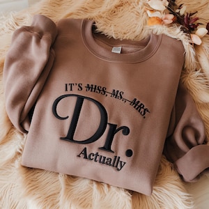 May include: A tan sweatshirt with the embroidered text "IT'S MISS, MS., MRS. Dr. Actually." The text is in black. The sweatshirt is folded on a fluffy, light brown surface.