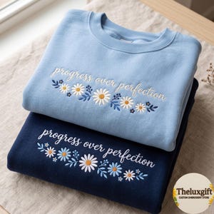May include: A light blue sweatshirt and a navy blue sweatshirt, both featuring white embroidered text that reads progress over perfection, accompanied by a floral design of white and yellow daisies with blue leaves and stems.