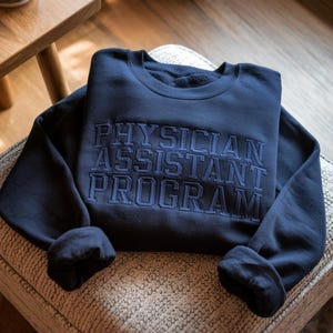 May include: A navy blue sweatshirt with the words "PHYSICIAN ASSISTANT PROGRAM" embroidered in a darker blue. The sweatshirt is folded neatly on a patterned surface, with the sleeves casually draped.